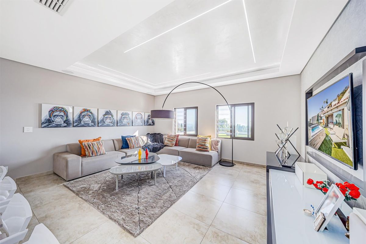 A modern, well-lit living room featuring a recessed ceiling with integrated LED lighting, large tiled floors, and aluminium-framed windows.