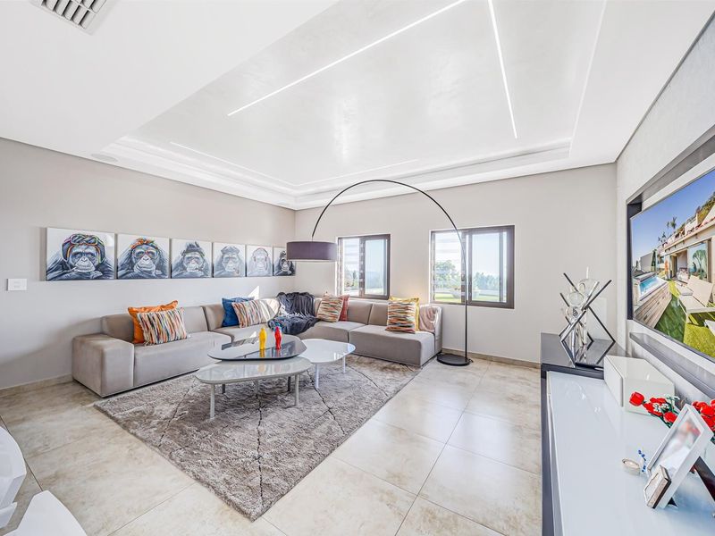 A modern, well-lit living room featuring a recessed ceiling with integrated LED lighting, large tiled floors, and aluminium-framed windows.