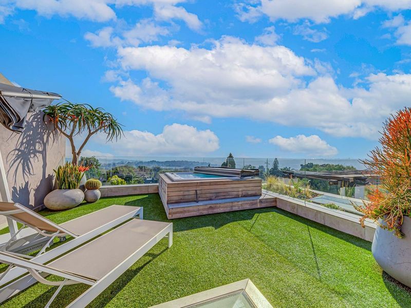 A modern rooftop terrace featuring a built-in spa, glass balustrades, and artificial turf, offering expansive elevated views.