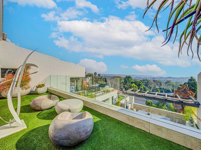 A modern elevated balcony featuring a glass balustrade, artificial turf, and a pergola, offering expansive views of the surrounding landscape.