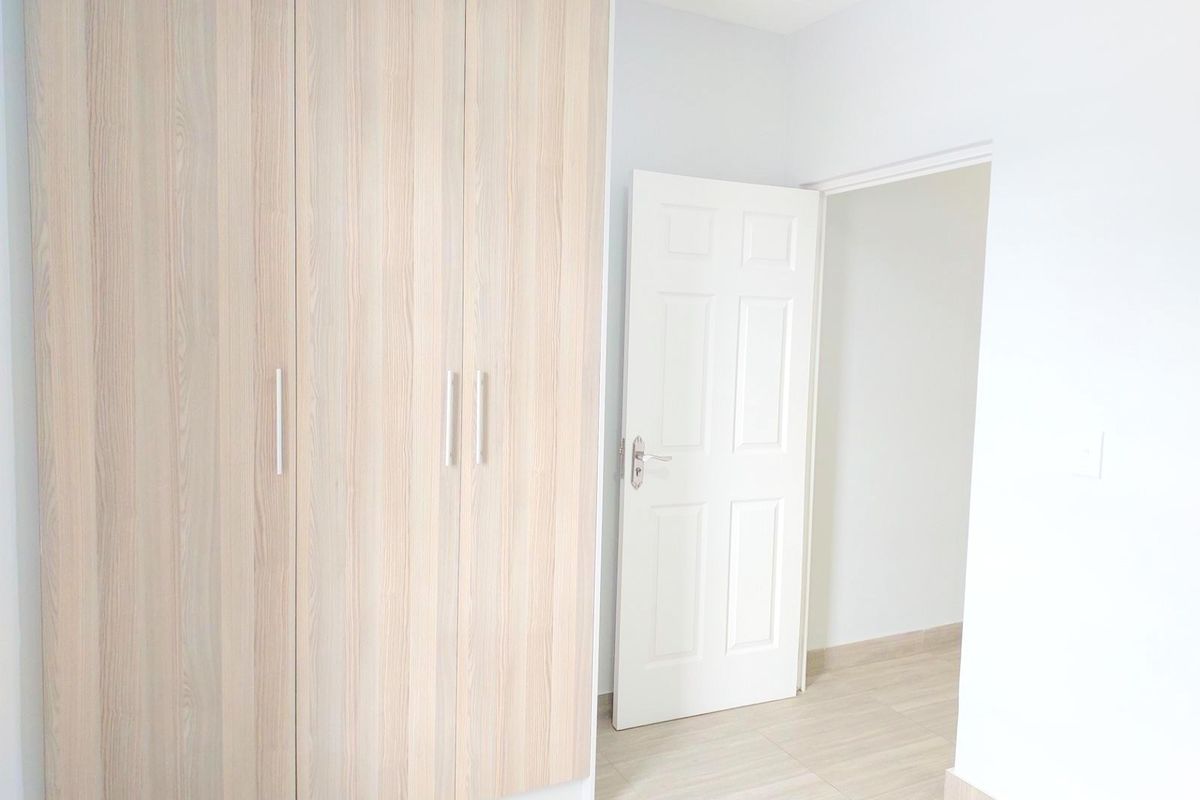A bright, empty bedroom featuring light wood-grain built-in wardrobes, neutral tiled flooring, and a clean white interior finish.