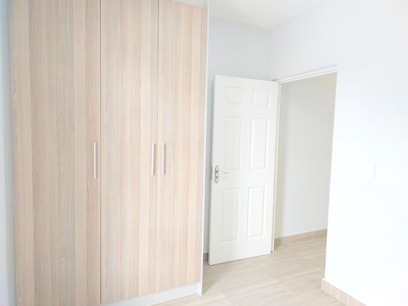A bright, empty bedroom featuring light wood-grain built-in wardrobes, neutral tiled flooring, and a clean white interior finish.