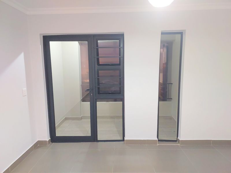 A bright, empty living area featuring neutral tiled floors, modern aluminium-framed glass doors, and a fixed glass panel providing access to an outdoor space.