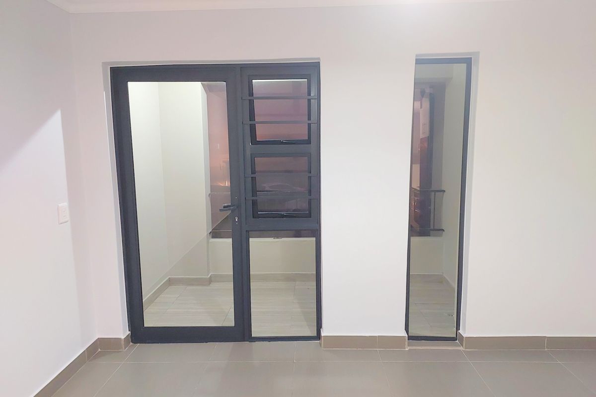 A bright, empty living area featuring neutral tiled floors, modern aluminium-framed glass doors, and a fixed glass panel providing access to an outdoor space.