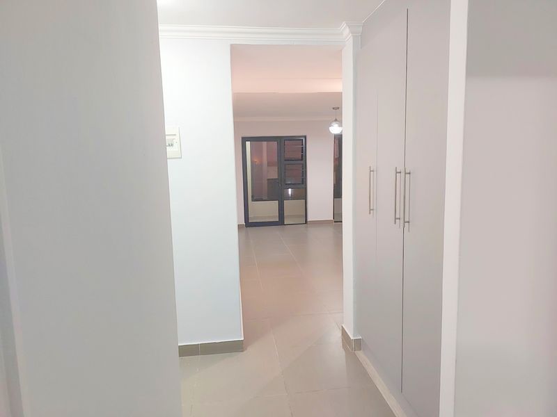 A clean, neutral hallway featuring built-in cupboards and tiled flooring, leading through to an open-plan living area.