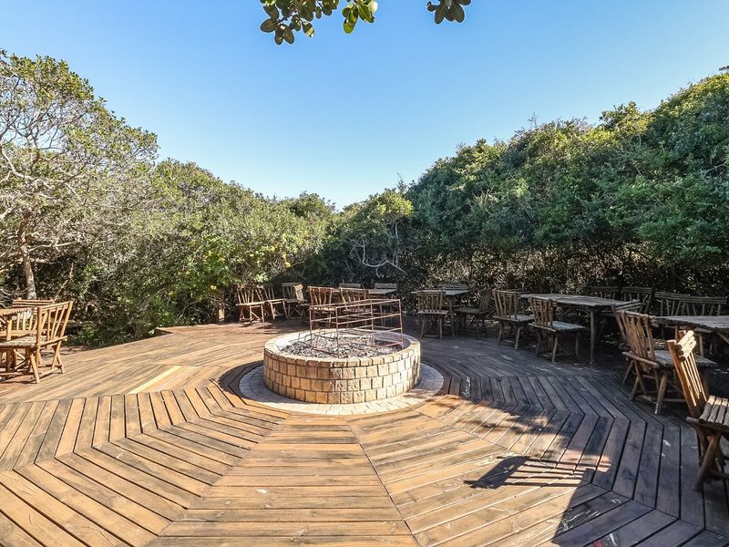 A spacious outdoor timber deck featuring a central circular brick fire pit, surrounded by mature natural vegetation.