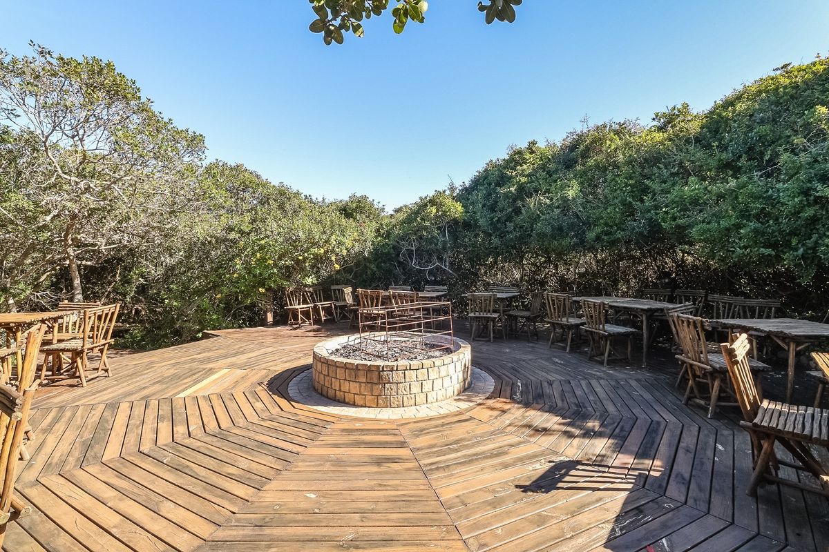 A spacious outdoor timber deck featuring a central circular brick fire pit, surrounded by mature natural vegetation.