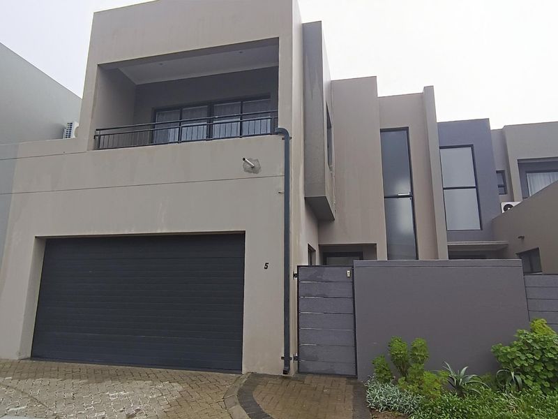 Modern multi-story townhouse facade featuring a double garage, private balcony with metal railing, and a paved driveway entrance.