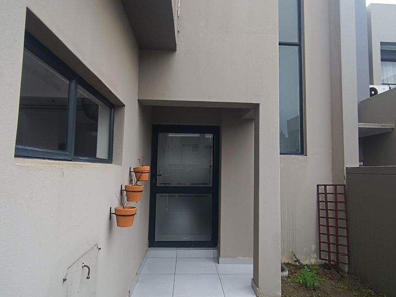 A modern rear exterior view featuring a tiled patio area, aluminium framed glass door, and large vertical windows with a neutral rendered finish.