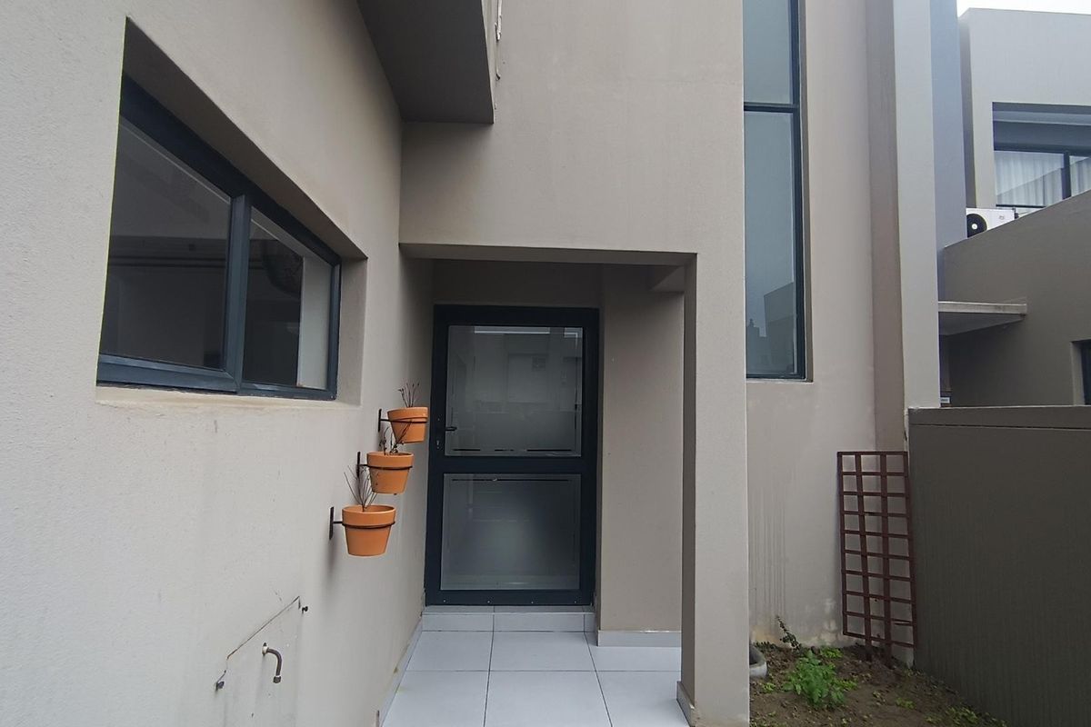 A modern rear exterior view featuring a tiled patio area, aluminium framed glass door, and large vertical windows with a neutral rendered finish.