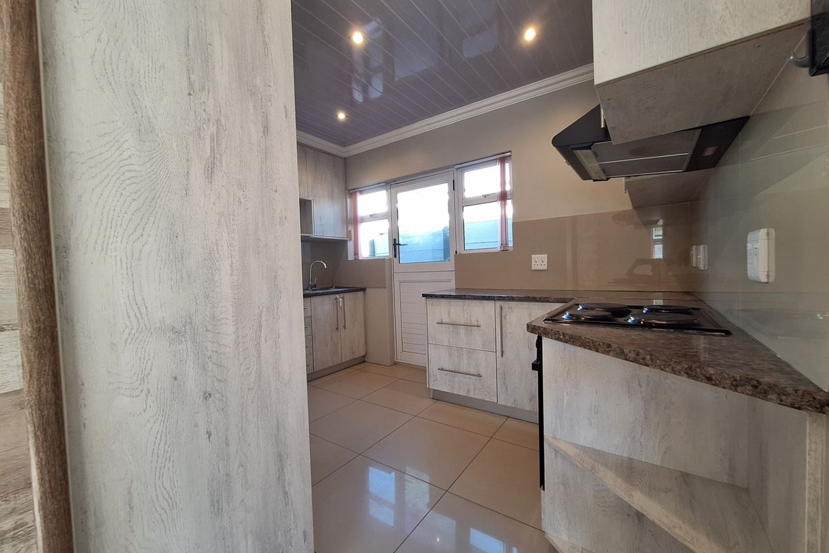 A clean, modern kitchen featuring light wood-grain cabinetry, granite-look countertops, an electric hob with extractor, and tiled flooring with direct exterior access.
