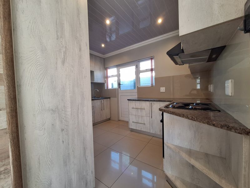 A clean, modern kitchen featuring light wood-grain cabinetry, granite-look countertops, an electric hob with extractor, and tiled flooring with direct exterior access.