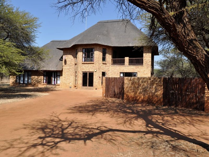 A two-story face brick house featuring a traditional thatch roof, a private balcony, and a secure brick boundary wall with a timber gate.