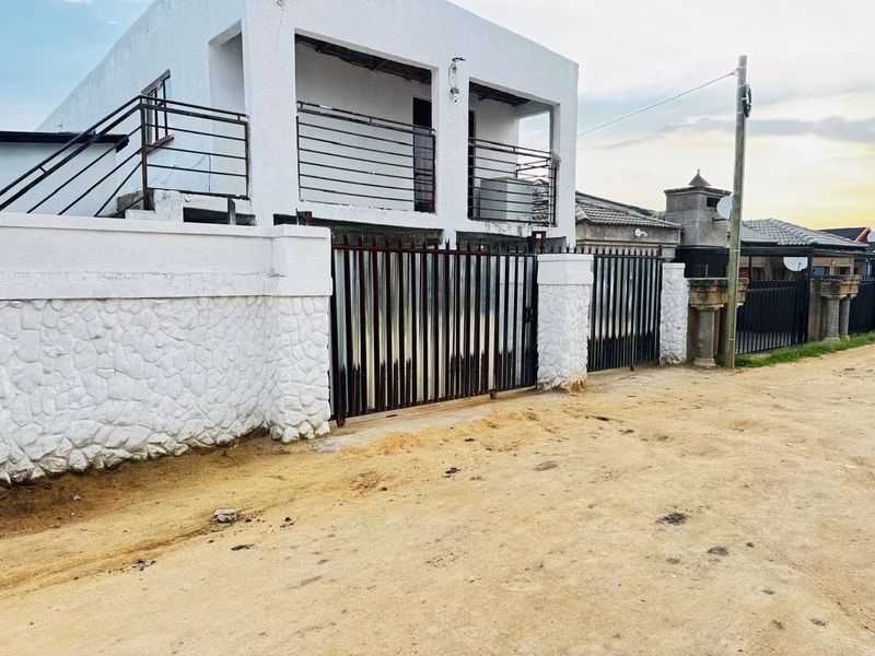 Front elevation of a two-story house featuring a metal security gate, stone-clad boundary wall, and upper-level balcony with metal railings.