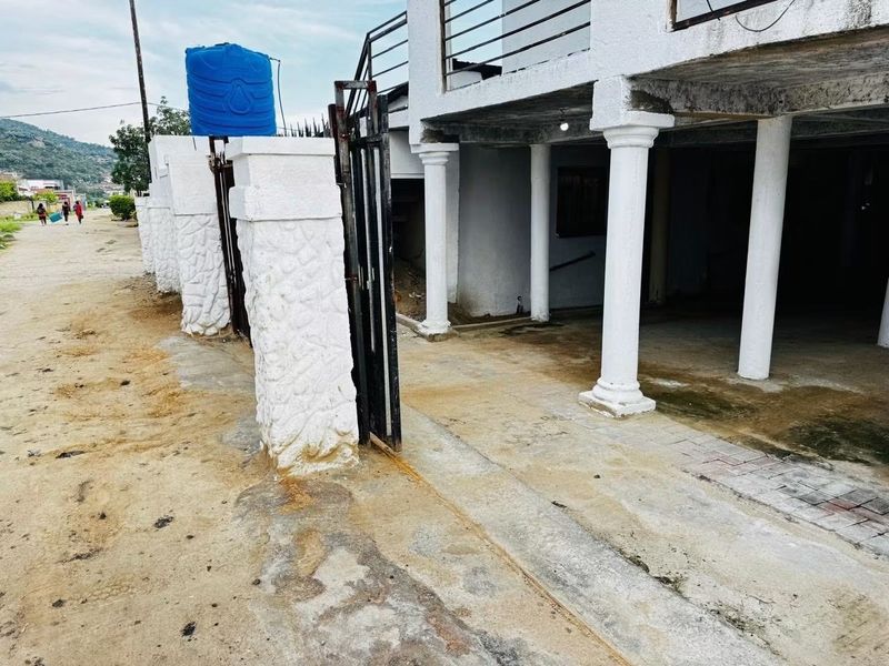 Front exterior view showing a driveway, stone-clad pillars, metal gate, and an elevated balcony area with a water storage tank.