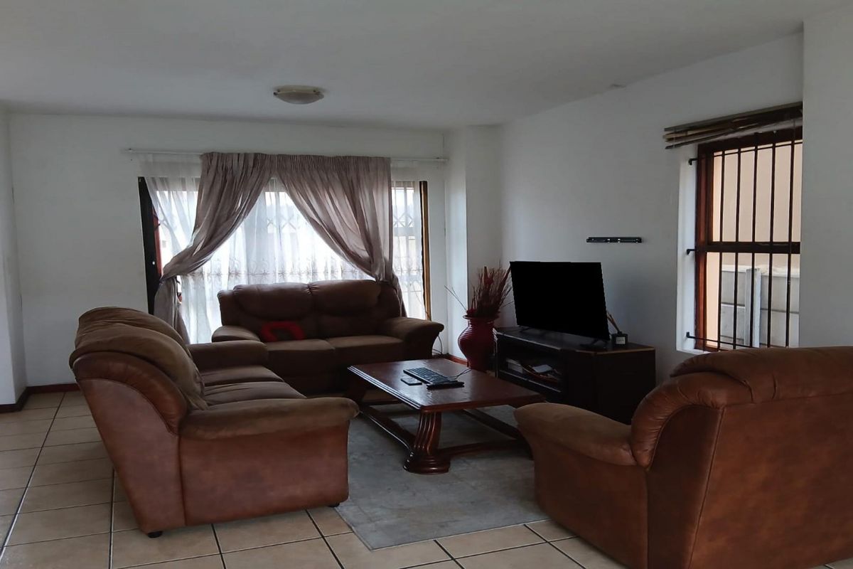 A spacious living room featuring neutral tiled flooring, large windows with security bars, and ample natural light.