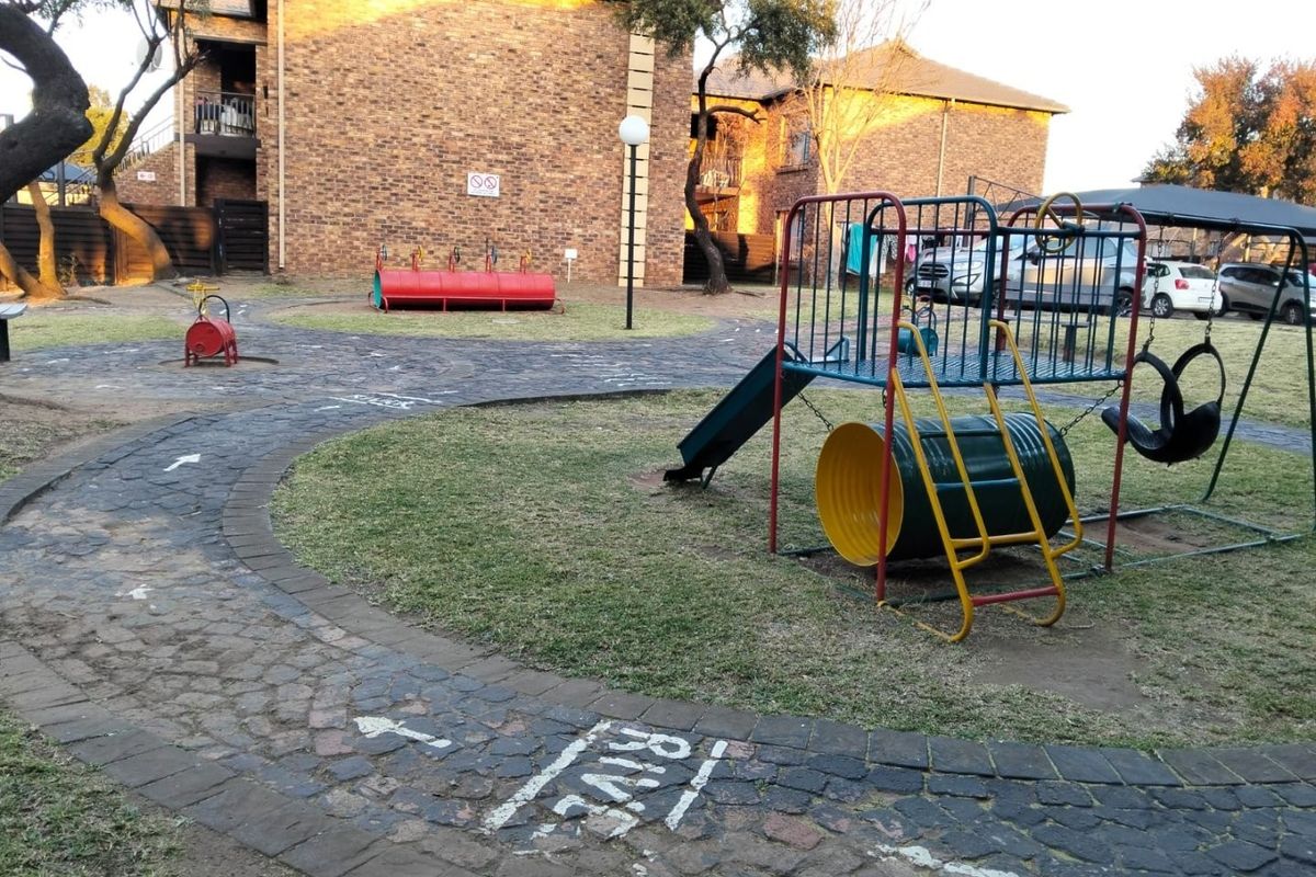 Communal outdoor area featuring a brick apartment building facade, paved pathways, and a playground set within a landscaped garden space.