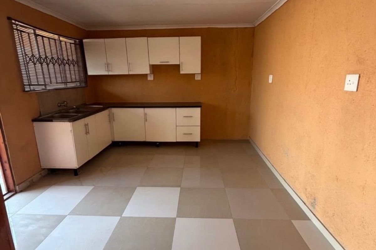 A compact, empty kitchen featuring white built-in cabinetry, a stainless steel sink, and a checkered tiled floor, requiring cosmetic updates to the wall finishes.