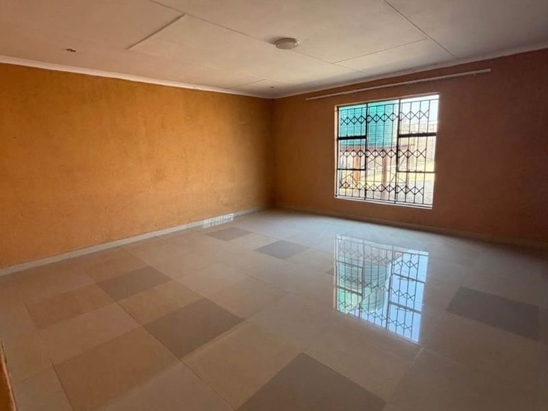 A spacious, empty living area featuring tiled flooring, orange-toned walls, and a window fitted with security bars.