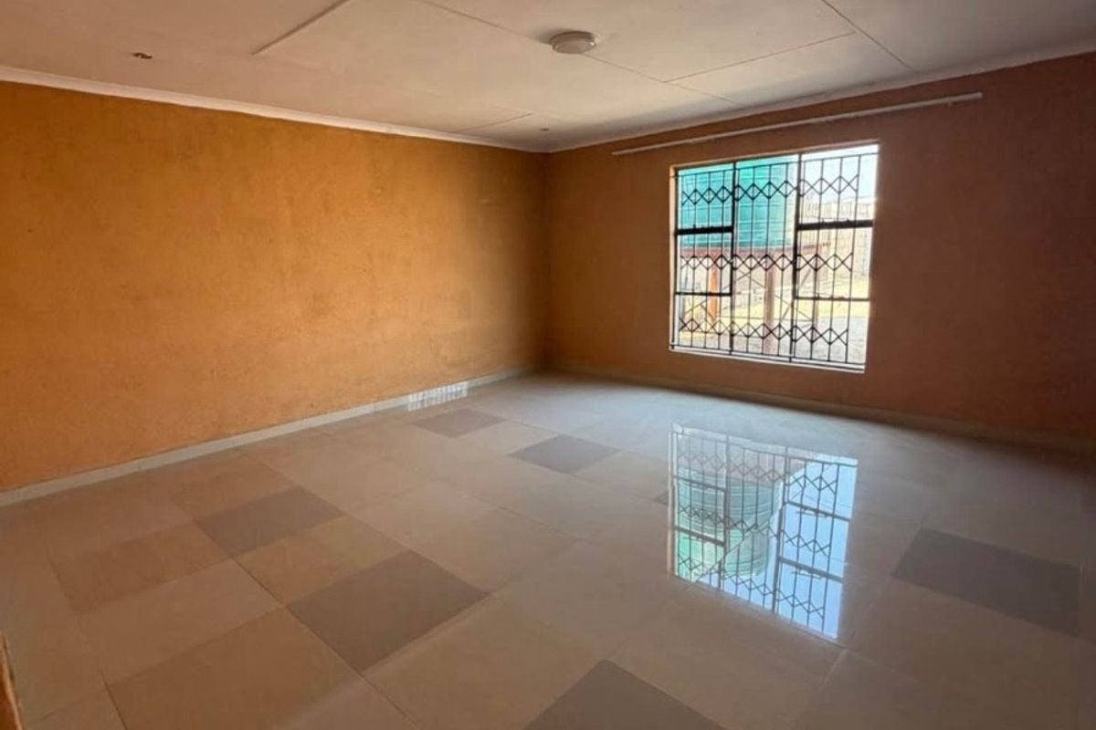 A spacious, empty living area featuring tiled flooring, orange-toned walls, and a window fitted with security bars.