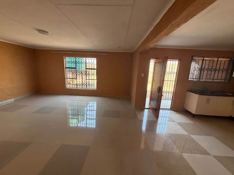 Spacious open-plan living area featuring tiled floors, neutral walls, and a compact kitchen corner with built-in cabinetry and sink.