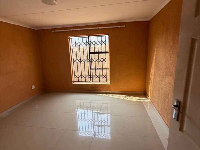 A bright, empty bedroom featuring neutral tiled flooring, textured orange walls, and a window fitted with a security gate.