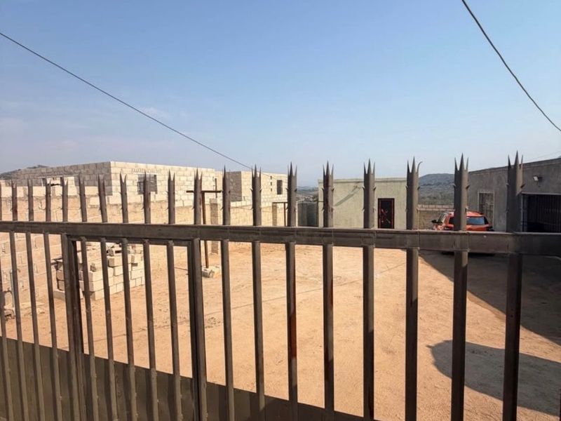 Front view of a property under construction, featuring a metal security gate, unpaved driveway, and unfinished masonry block structures.