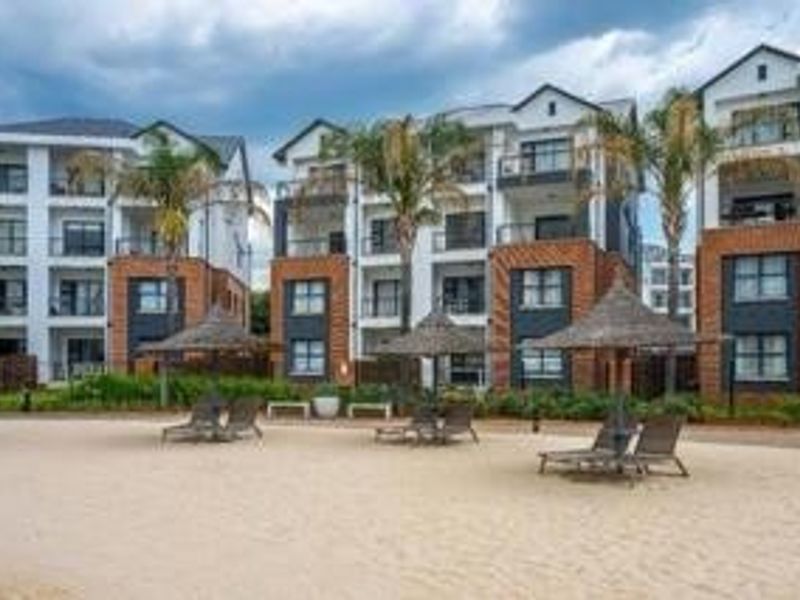 Exterior view of modern multi-story apartment buildings featuring brick facades, private balconies, and a communal sandy recreation area with thatched umbrellas.