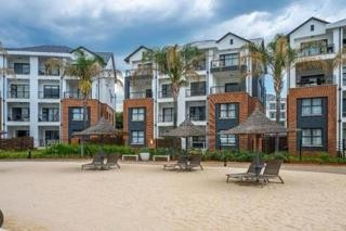 Exterior view of modern multi-story apartment buildings featuring brick facades, private balconies, and a communal sandy recreation area with thatched umbrellas.