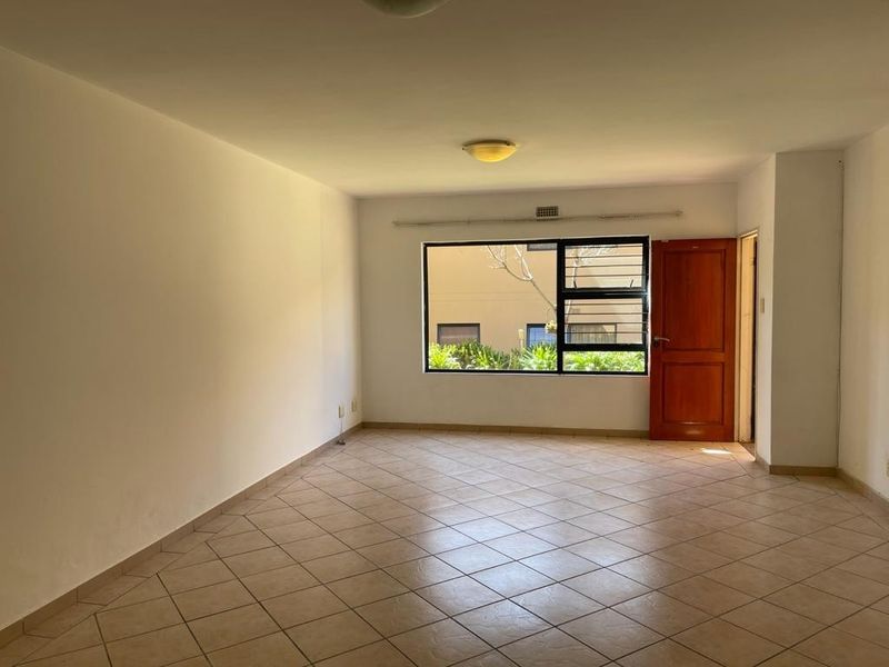 A spacious, empty living room featuring neutral tiled flooring, a large window with security bars, and a wooden entrance door.