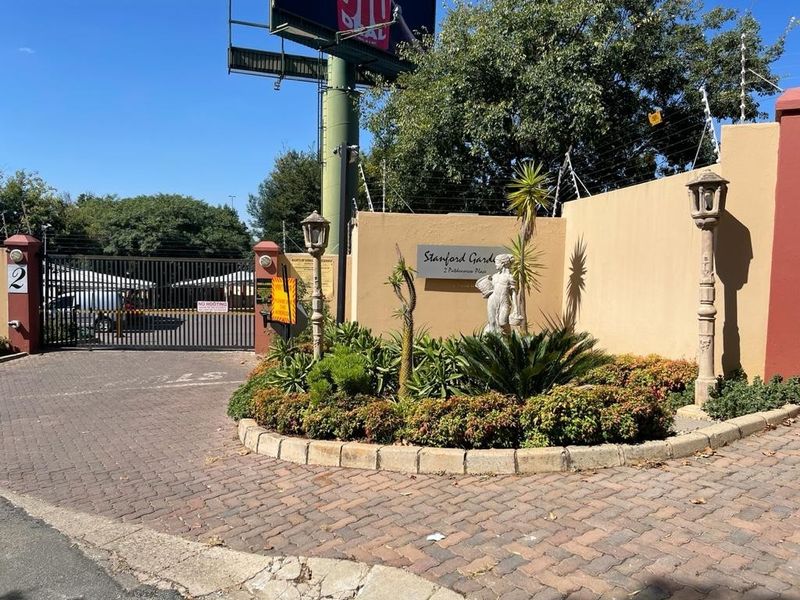 Entrance to Stanford Gardens townhouse complex featuring a paved driveway, security gate, and perimeter boundary wall with electric fencing.
