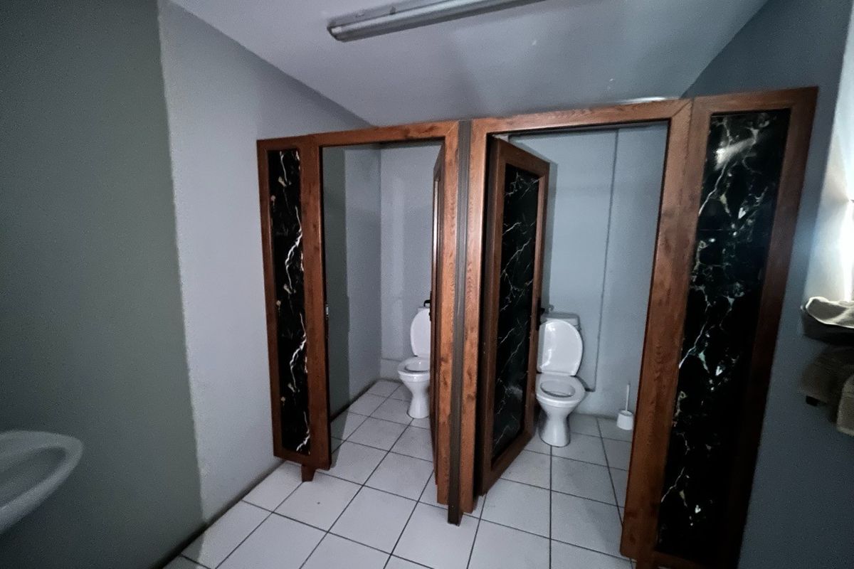 A functional industrial-style bathroom featuring two toilet cubicles with wooden frames and marble-effect accents, finished with white floor tiles.