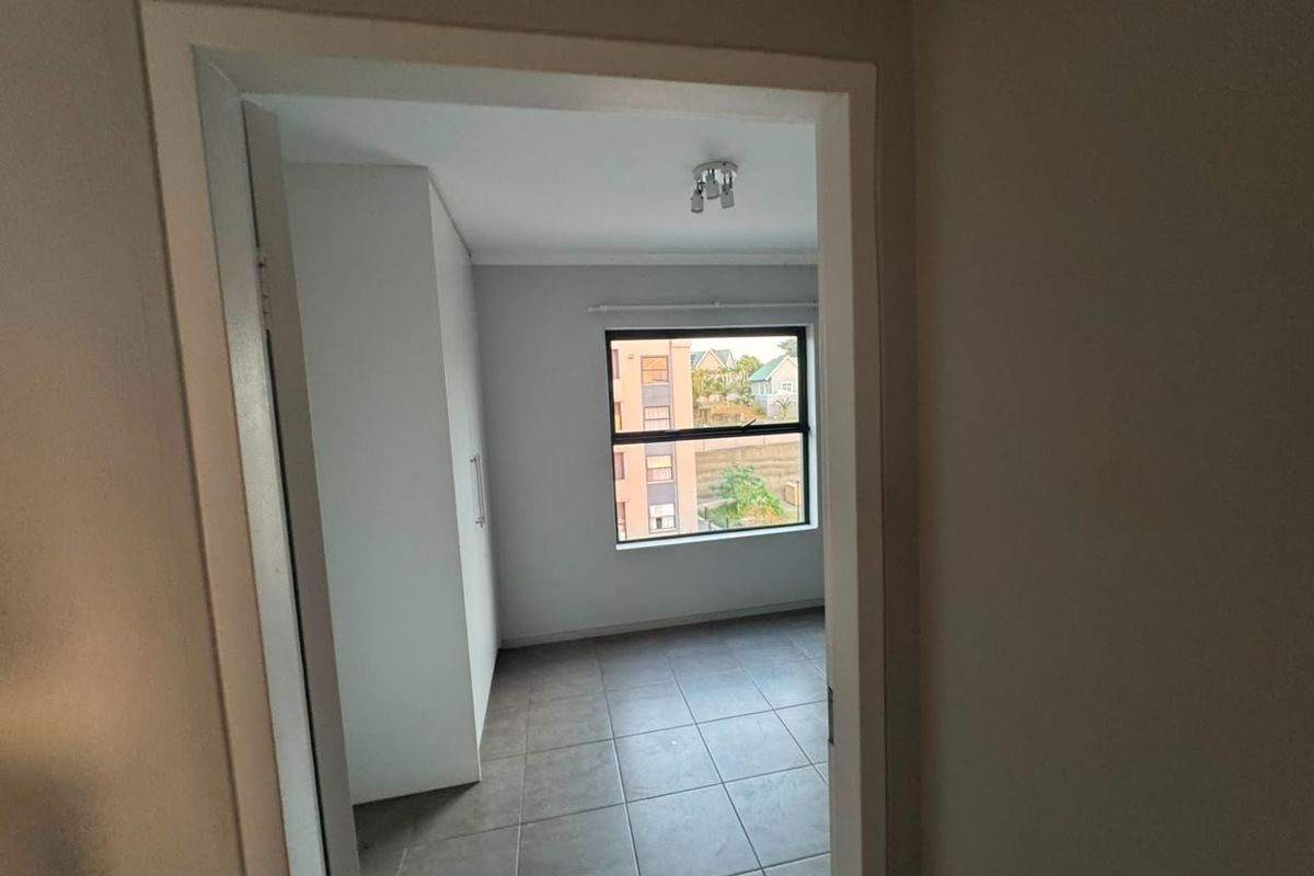 A bright, empty bedroom featuring neutral tiled flooring, a built-in wardrobe, and a window with views of the surrounding complex.