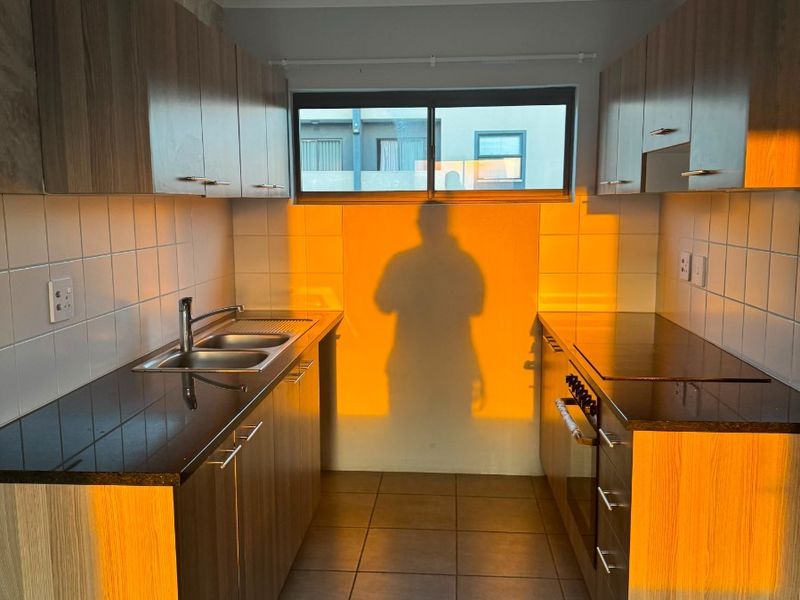 A functional galley-style kitchen featuring wood-finish cabinetry, dark countertops, a double sink, and built-in oven, illuminated by warm natural light.
