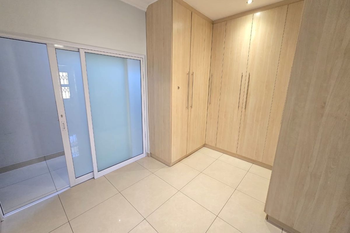 A bright, empty bedroom featuring modern light wood built-in cupboards, neutral tiled flooring, and a sliding glass door providing access to an adjacent space.