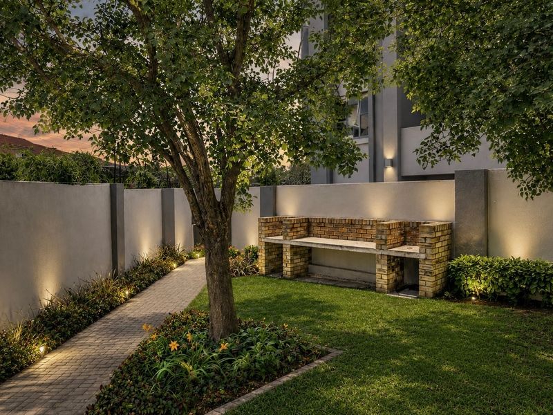 A well-maintained garden featuring a built-in brick braai, paved walkway, lush lawn, and modern perimeter wall lighting.