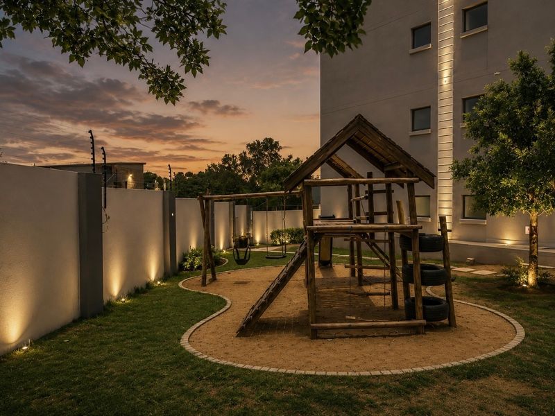 A well-maintained communal garden area featuring a wooden playground structure, manicured lawn, and integrated architectural lighting against a modern building facade.