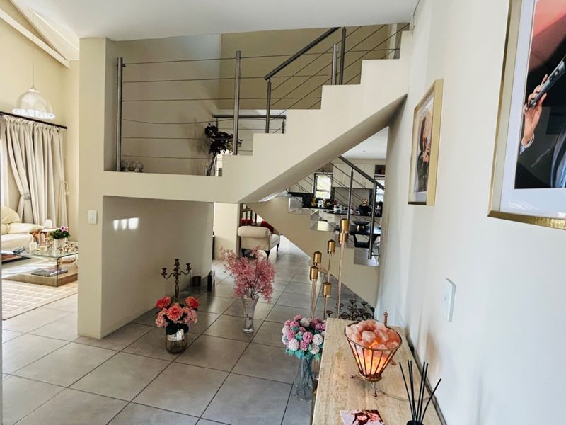 A bright entrance hallway featuring a modern staircase with stainless steel balustrades and neutral tiled flooring, leading into an open-plan living area.