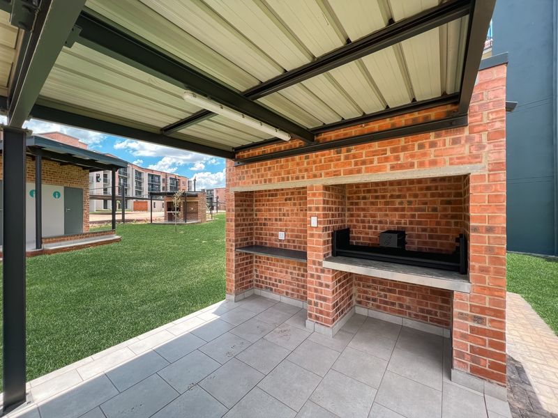 A covered outdoor patio area featuring a built-in brick braai, tiled flooring, and a metal roof structure, overlooking a communal garden space.