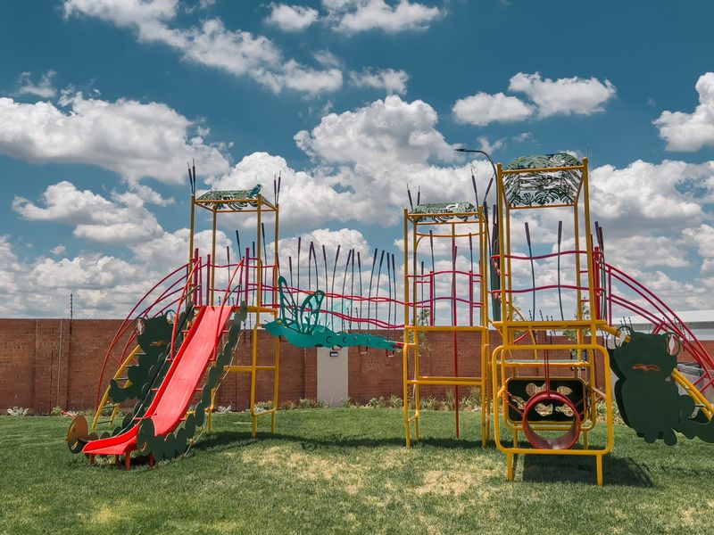 A well-maintained communal garden area featuring a colorful children's playground structure, set against a secure brick boundary wall with electric fencing.