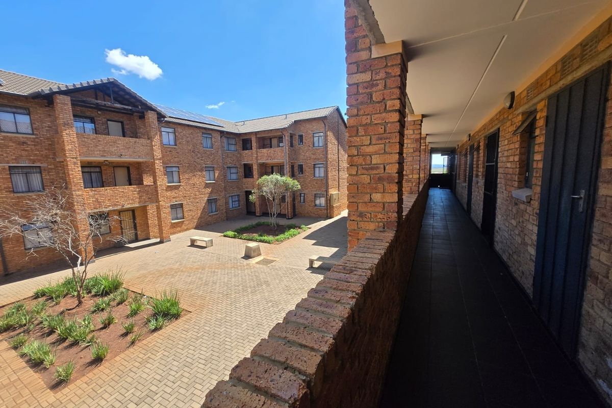 View of an apartment complex courtyard featuring a paved common area, landscaped garden beds, and a covered exterior walkway with brick finishes.