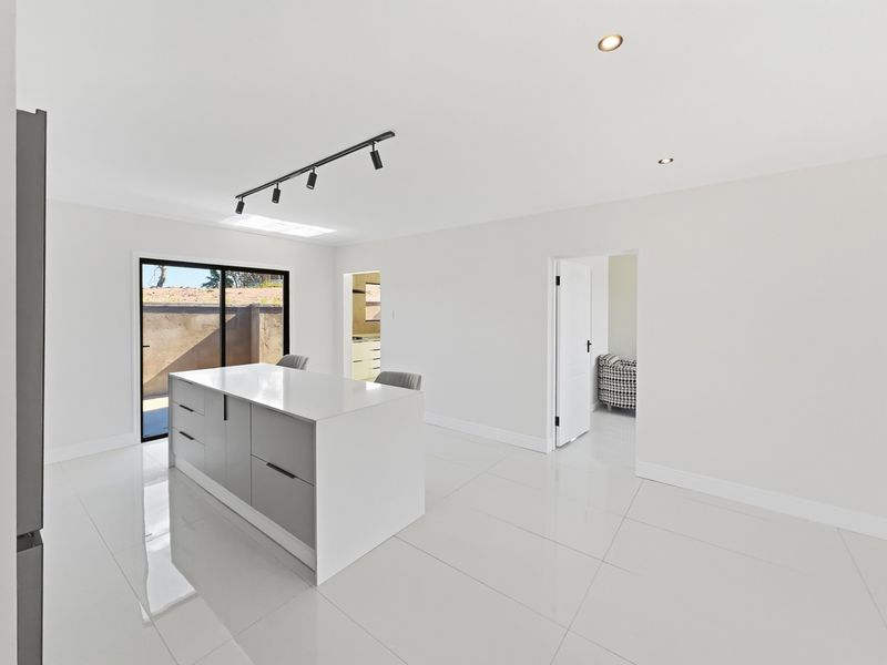 Modern, minimalist kitchen featuring a central island, sleek cabinetry, polished floor tiles, and abundant natural light from a skylight and sliding glass doors.