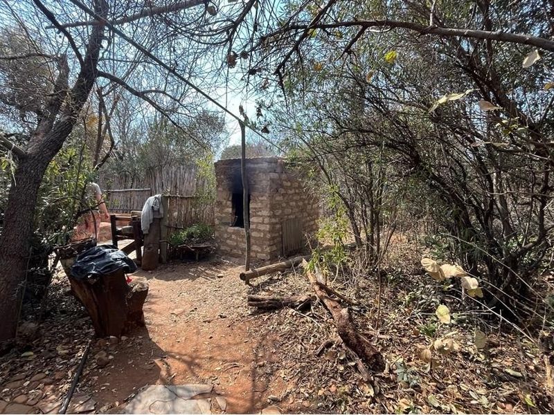 A rustic outdoor area featuring a stone-built oven and reed fencing, situated within a natural, wooded farm setting.