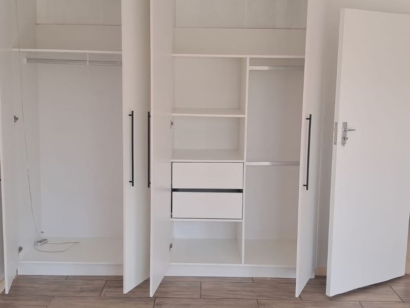 A clean, empty bedroom featuring modern white built-in cupboards with integrated shelving and drawers, complemented by neutral tiled flooring.