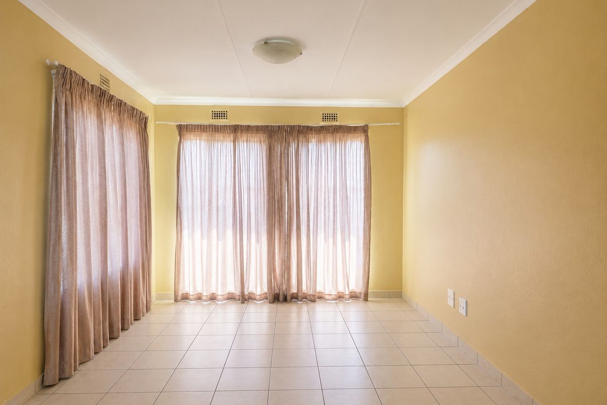 A bright, empty living room featuring neutral tiled flooring, yellow walls, and large windows with curtains.