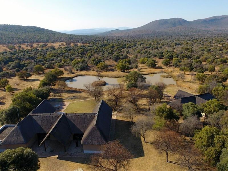 Aerial view of a farm property featuring multiple thatched-roof buildings, a natural dam, and expansive surrounding bushveld landscape.