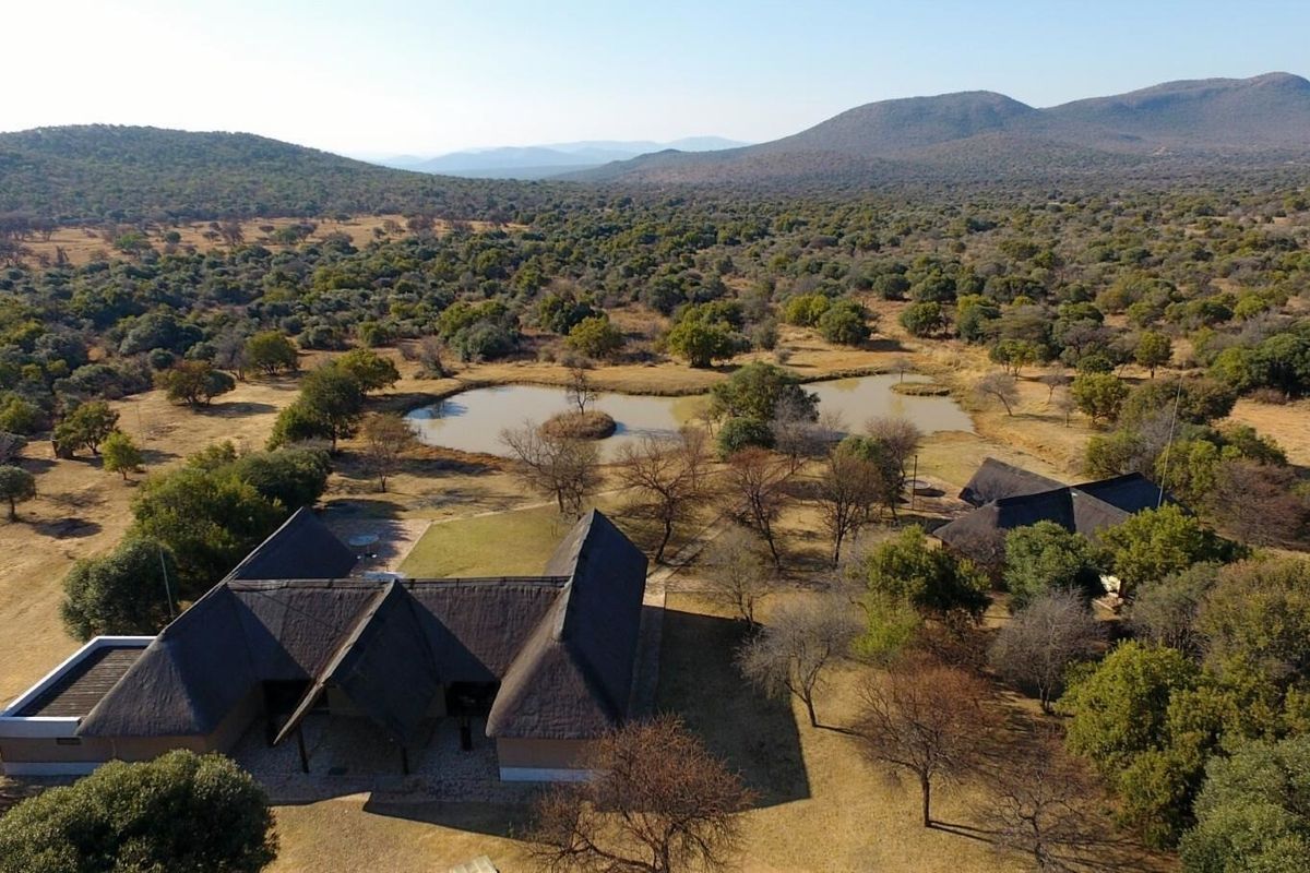 Aerial view of a farm property featuring multiple thatched-roof buildings, a natural dam, and expansive surrounding bushveld landscape.