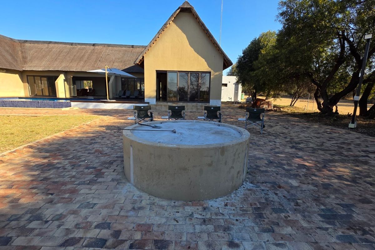 Rear exterior view of a thatched-roof farm property featuring a paved patio, built-in fire pit, and an adjacent swimming pool.