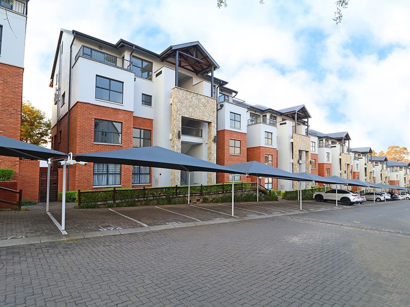 Front elevation of a multi-story apartment complex featuring brick and stone facade, balconies, and covered parking bays with shade cloth structures.