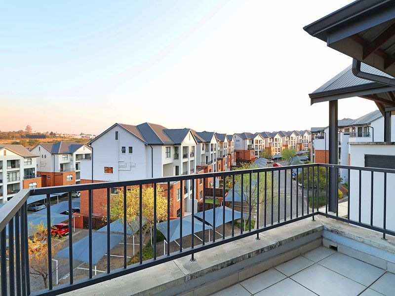A spacious balcony featuring tiled flooring and a metal balustrade, offering an elevated view of the surrounding apartment complex and parking area.